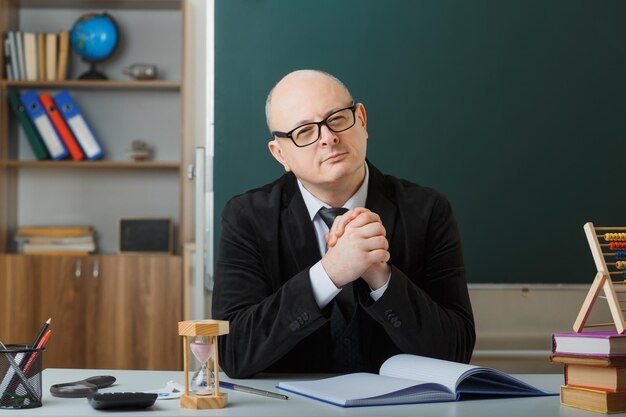 Mann, Lehrer, der eine Brille trägt, die das Klassenbuch überprüft und in die Kamera blickt, die Augen zusammenkneift, skeptisch lächelt und an der Schulbank vor der Tafel im Klassenzimmer sitzt