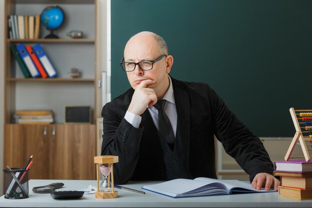 Mann, Lehrer, der eine Brille trägt, die das Klassenbuch überprüft und mit nachdenklichem Gesichtsausdruck zur Seite schaut und die Hand am Kinn hält, sitzt an der Schulbank vor der Tafel im Klassenzimmer