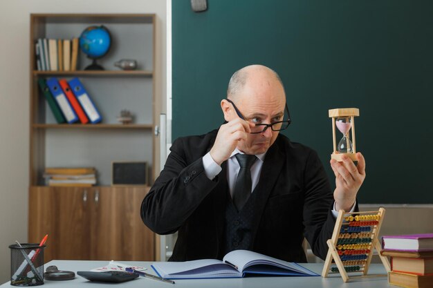 Mann, Lehrer, der eine Brille trägt, sitzt an der Schulbank vor der Tafel im Klassenzimmer und hält eine Sanduhr, die den Unterricht erklärt und fasziniert aussieht