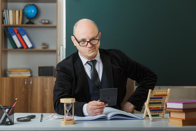 Mann Lehrer mit Brille sitzt an der Schulbank mit Klassenbuch vor der Tafel im Klassenzimmer und erklärt den Unterricht, der auf den Taschenrechner schaut, der unzufrieden ist
