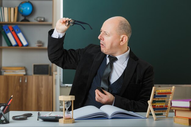 Mann Lehrer mit Brille sitzt an der Schulbank mit Klassenbuch vor der Tafel im Klassenzimmer und erklärt den Unterricht selbstbewusst