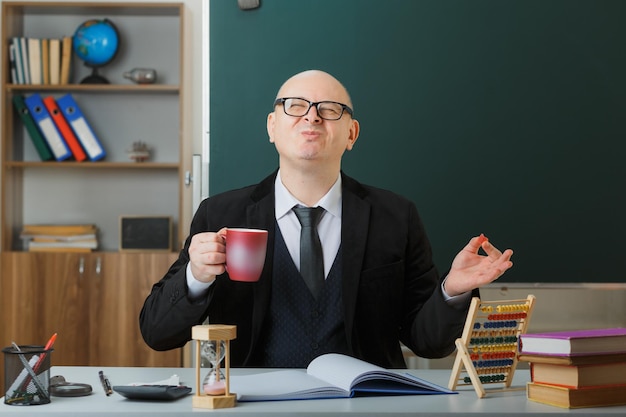 Mann Lehrer mit Brille sitzt an der Schulbank vor der Tafel im Klassenzimmer und hält eine Tasse Kaffee glücklich und erfreut lächelnd