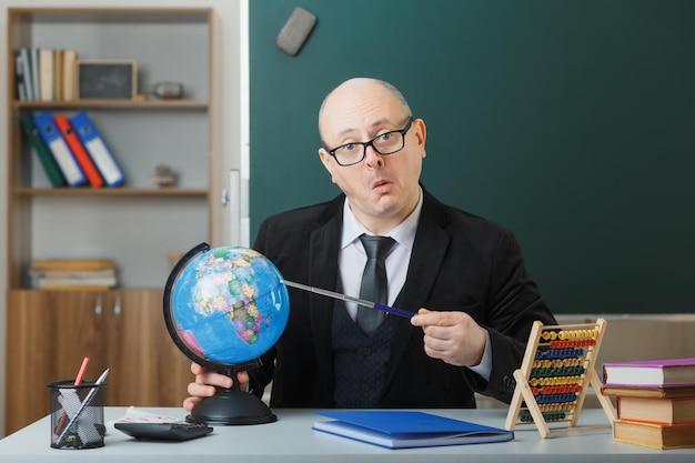 Mann Lehrer mit Brille sitzt mit Globus an der Schulbank vor der Tafel im Klassenzimmer und erklärt den Unterricht und sieht verwirrt aus
