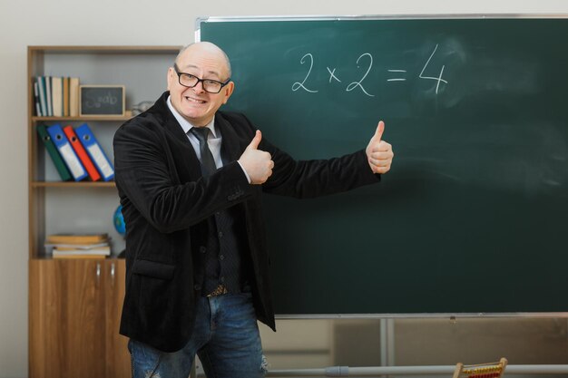 Mann Lehrer mit Brille steht in der Nähe der Tafel im Klassenzimmer und erklärt den Unterricht, der Daumen hoch zeigt, glücklich und zufrieden