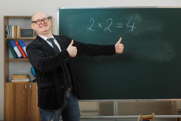 Mann Lehrer mit Brille steht in der Nähe der Tafel im Klassenzimmer und erklärt den Unterricht, der Daumen nach oben zeigt, glücklich und erfreut, breit lächelnd