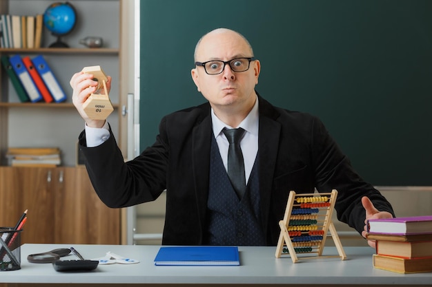 Mann Lehrer mit Brille und Klassenbuch sitzt an der Schulbank vor der Tafel im Klassenzimmer und schwingt Sanduhr wütend und genervt