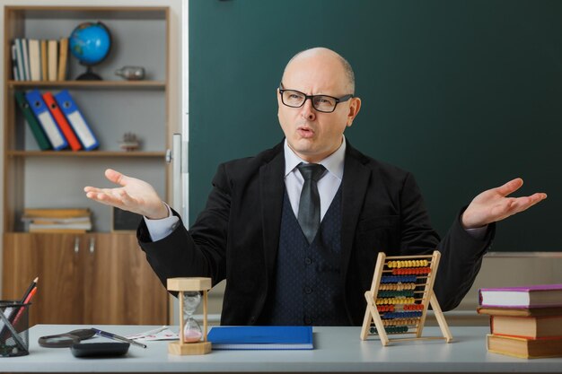 Mann Lehrer mit Brille und Klassenbuch sitzt an der Schulbank vor der Tafel im Klassenzimmer und sieht unzufrieden aus, die Arme vor Unmut hebend