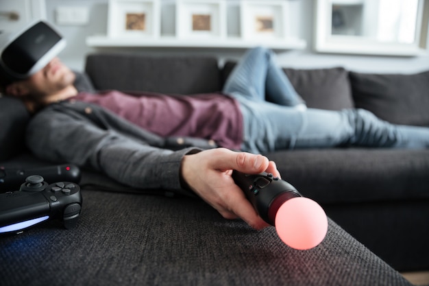 Mann liegt auf Sofa und trägt eine Virtual-Reality-Brille mit Joystick.