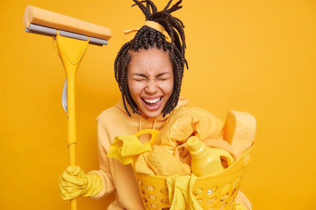 Mann macht Hausarbeiten hält Mop zum Waschen des Bodens im Haus trägt einen Korb mit Wäschekorb mit Reinigungsmitteln isoliert auf Gelb