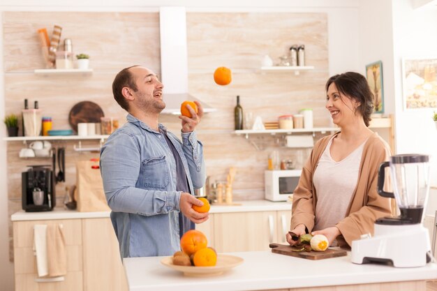 Mann macht Tricks mit Orangen für die Frau in der Küche, während er einen gesunden Smoothie zubereitet. Gesunder, unbeschwerter und fröhlicher Lebensstil, Ernährung und Frühstückszubereitung am gemütlichen sonnigen Morgen