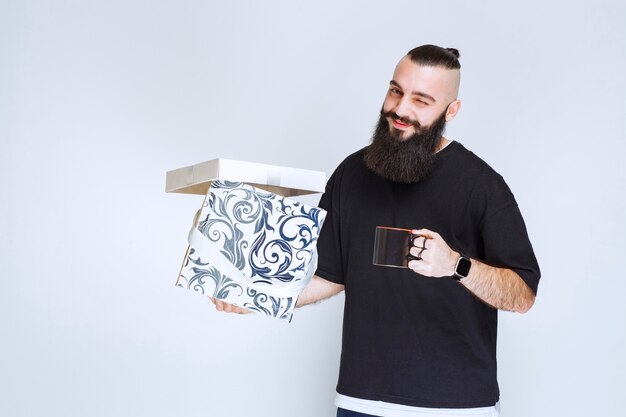 Mann mit Bart, der eine offene blaue Geschenkbox mit einer Tasse Kaffee hält und glücklich aussieht.