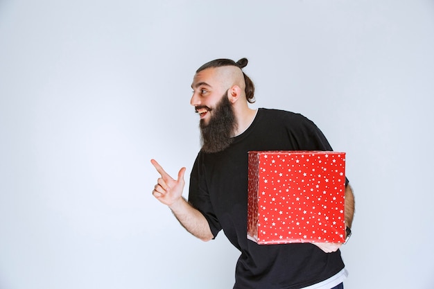 Mann mit Bart, der eine rote Geschenkbox hält und jemandem zeigt.