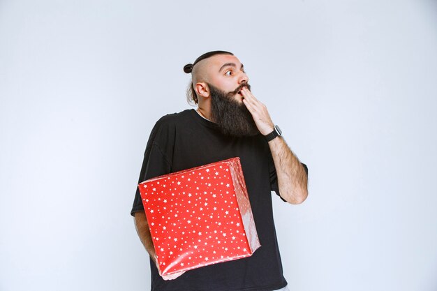 Mann mit Bart, der eine rote Geschenkbox hält und verwirrt und verängstigt aussieht.