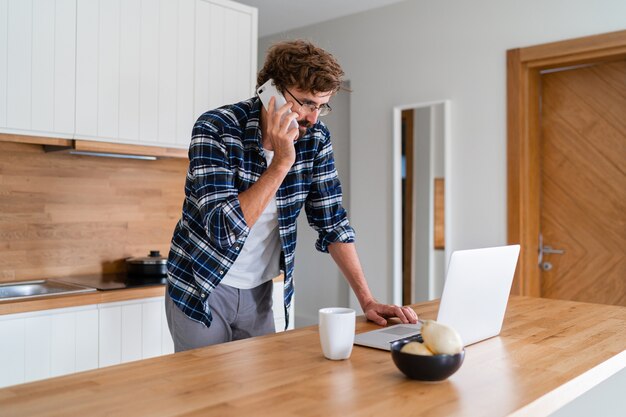 Mann mit Bart, der per Telefon spricht und Laptop auf der Küche benutzt.