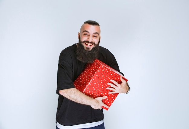 Mann mit Bart, der seine rote Geschenkbox hält, sie genießt und sich glücklich fühlt.