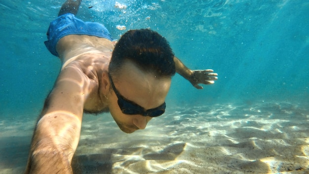 Kostenloses Foto mann mit brille, der unter dem blauen und transparenten wasser des mittelmeers schwimmt. halten der kamera