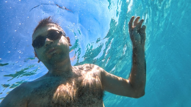 Mann mit Brille, der unter dem blauen und transparenten Wasser des Mittelmeers schwimmt. Halten der Kamera