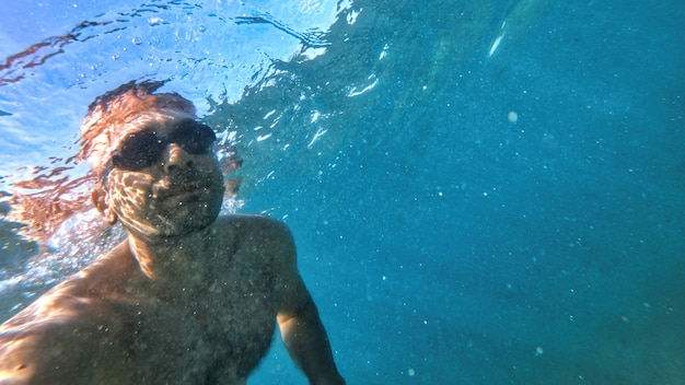 Mann mit Brille, der unter dem blauen und transparenten Wasser des Mittelmeers schwimmt. Halten der Kamera