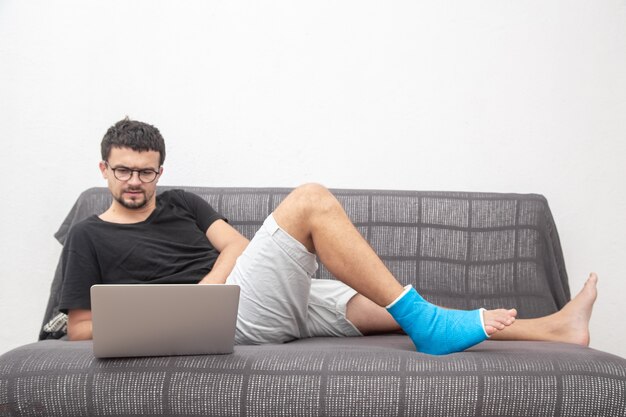 Mann mit Brille mit gebrochenem Bein in blauer Schiene zur Behandlung von Verletzungen durch Knöchelverstauchung, die an einem Laptop auf Couch zu Hause arbeiten.