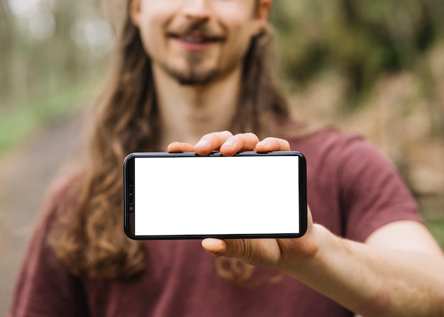 Mann mit dem langen Haar, das Smartphoneschablone in der Natur zeigt