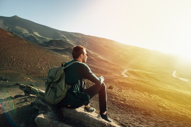 Mann mit dem Rucksack, der auf Felsen bei Sonnenuntergang auf Vulkan Ätna-Berg in Sizilien sitzt