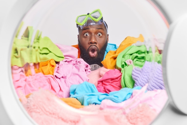 Mann mit dickem Bart trägt eine Schnorchelmaske auf einem Haufen, umgeben von bunten schmutzigen Kleidern in der Waschmaschine lädt die Waschmaschine die Hausarbeit