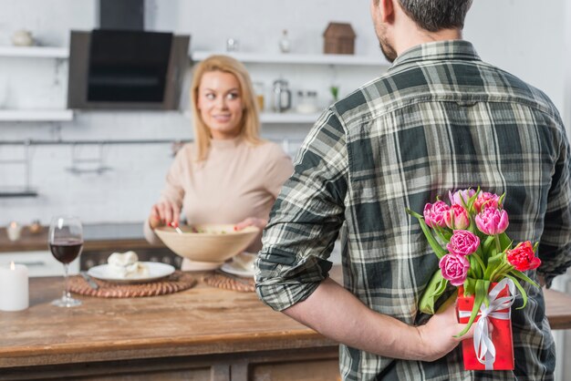 Mann mit Geschenk und Blumen von hinten und Frau in der Küche