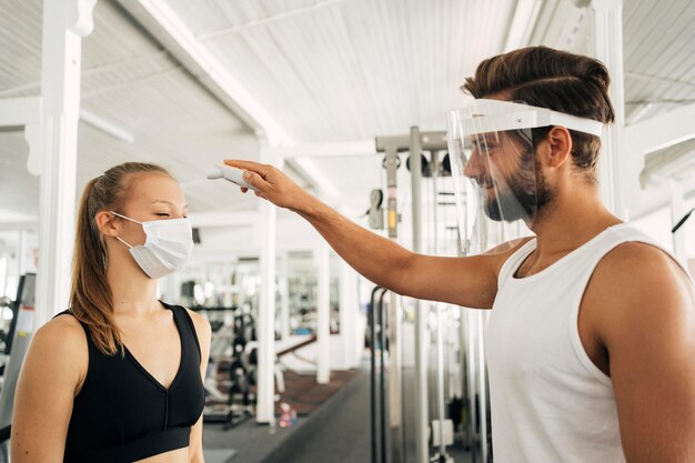 Mann mit Gesichtsschutz prüft die Temperatur der Frau im Fitnessstudio