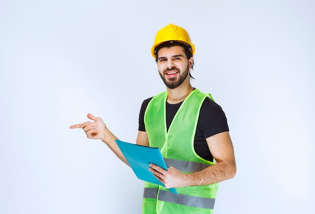 Mann mit Helm, der einen blauen Ordner hält und auf jemanden auf der linken Seite zeigt.