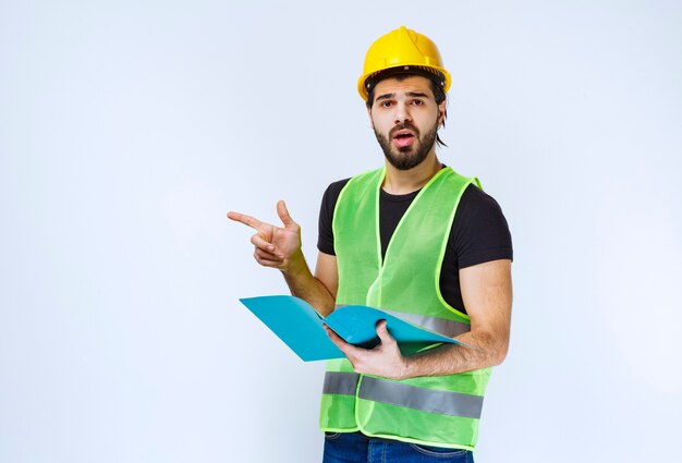 Mann mit Helm, der einen blauen Ordner hält und auf jemanden auf der linken Seite zeigt.