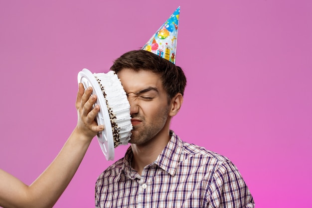 Mann mit Kuchen auf Gesicht über lila Wand. Geburtstagsfeier.