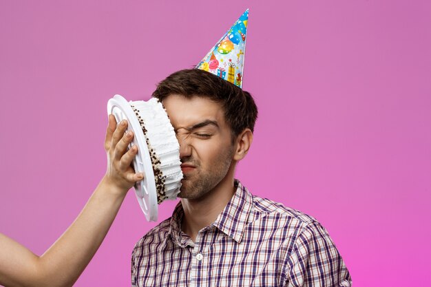 Mann mit Kuchen auf Gesicht über lila Wand. Geburtstagsfeier.