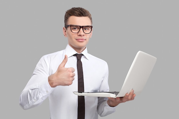 Mann mit laptop. hübscher junger mann in hemd und krawatte, der einen laptop hält und gestikuliert, während er vor grauem hintergrund steht