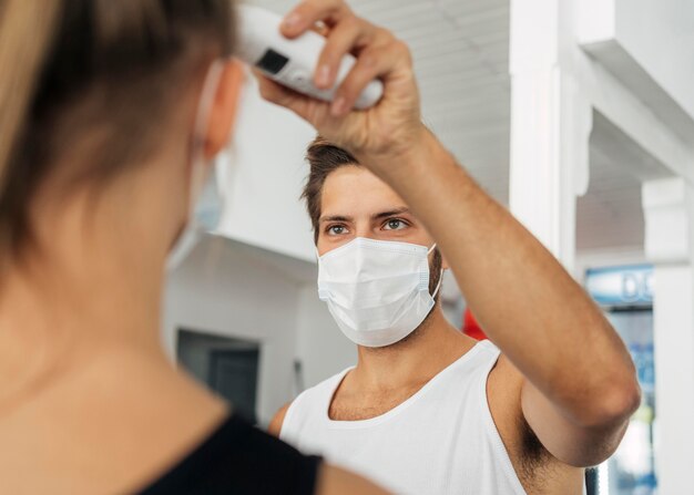 Mann mit medizinischer Maske im Fitnessstudio, das die Temperatur der Frau prüft