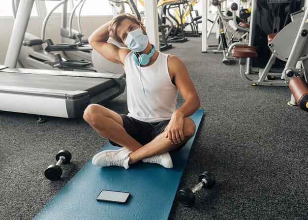 Mann mit medizinischer Maske und Kopfhörern im Fitnessstudio, das auf Matte trainiert