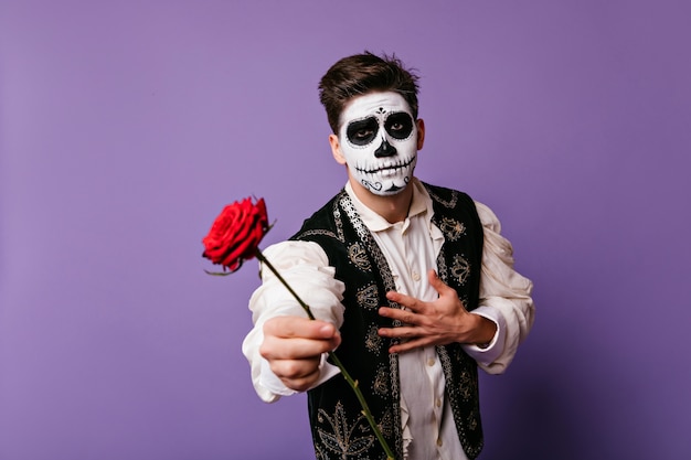 Mann mit mexikanischem toten Make-up, das rote Blume hält. emotionaler Typ in traditioneller spanischer Kleidung.