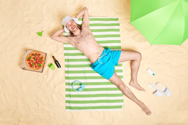 Mann mit nacktem Oberkörper lächelt glücklich trägt Sonnenhut und blaue Shorts posiert oben ohne auf gestreiftem Handtuch umgeben von Strandaccessoires hat faulen Tag gute Erholung am Meer. Sommerzeitkonzept