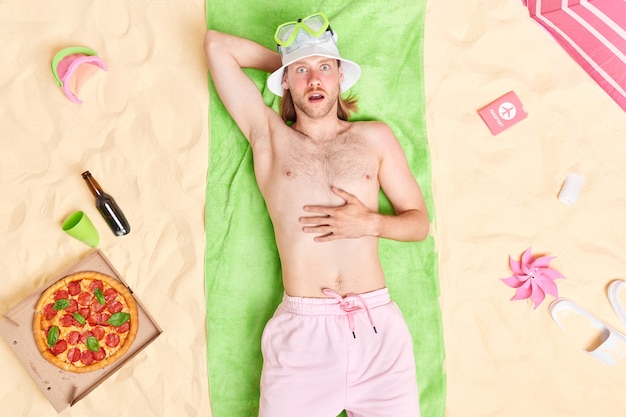 Mann mit nacktem oberkörper rotes sonnenverbranntes gesicht liegt auf grünem handtuch verbringt sommerurlaub am meer isst pizza trinkt bier posiert am sandstrand trägt panama und shorts