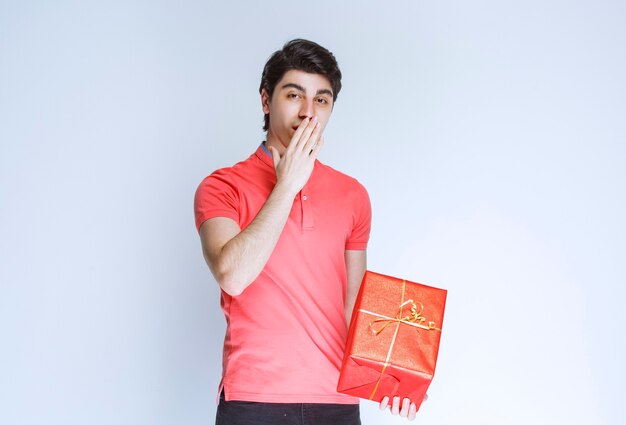 Mann mit roter Geschenkbox, die Hand an seinen Mund legt und denkt.