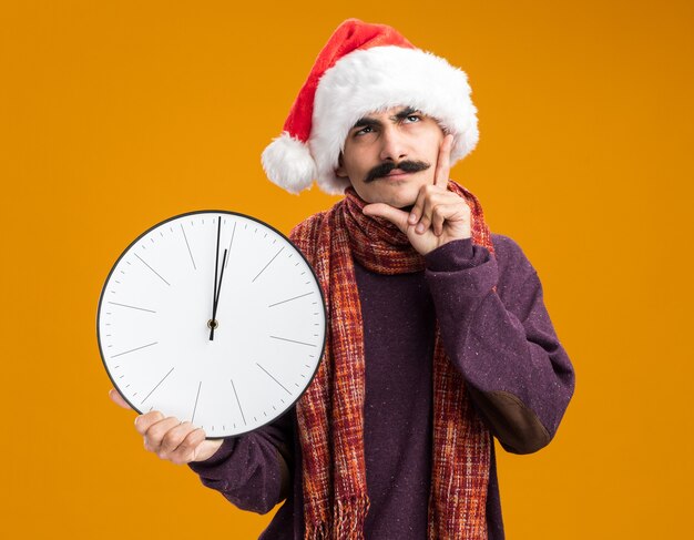 Mann mit Schnurrbart, der eine Weihnachtsmütze mit einem warmen Schal um den Hals trägt und eine Uhr hält, die verwirrt über der orangefarbenen Wand steht?