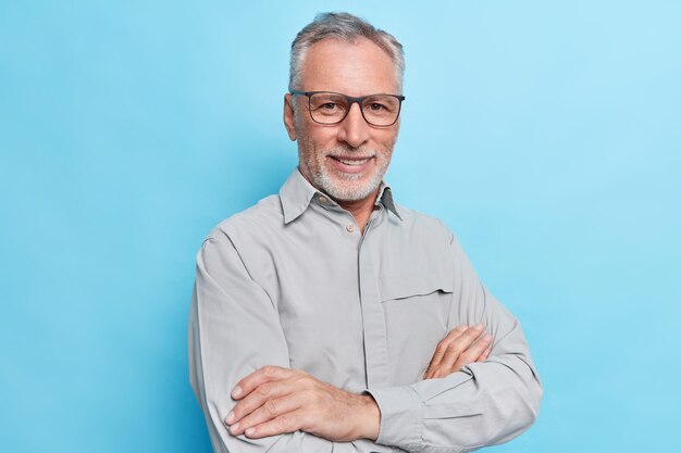 Mann mit verschränkten Armen, selbstbewusstem, fröhlichem Gesichtsausdruck, formellem Hemd und Brille zur Sehkorrektur vor blauer Wand