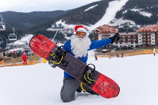 Mann mit weihnachtsmann-hut mit einem snowboard in einem skigebiet.