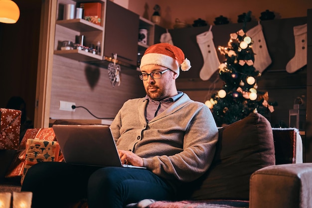 Mann mit Weihnachtsmütze und Brille in warmem Pullover sitzt auf dem Sofa und arbeitet zu Weihnachten am Laptop.