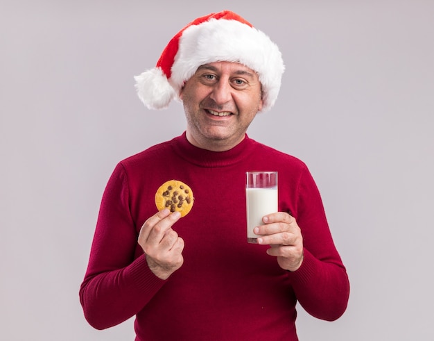 Kostenloses Foto mann mittleren alters, der eine weihnachtsmütze mit einem glas milch und keksen trägt und glücklich und fröhlich über weißer wand steht?