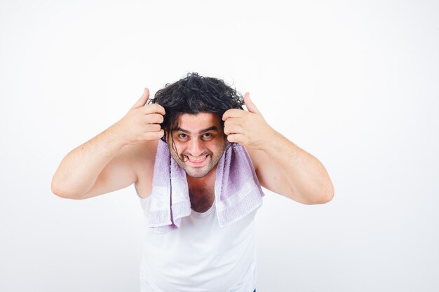 Mann mittleren Alters, der Haarsträhne hält, während Kamera in Tank Top, Handtuch und glücklich, Vorderansicht betrachtet.