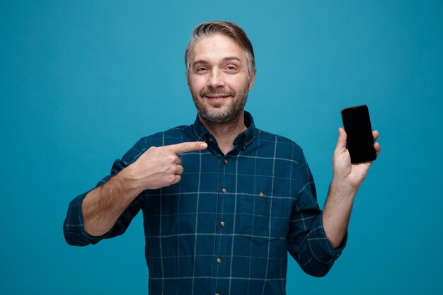 Mann mittleren Alters mit grauem Haar in dunklem Hemd, der das Smartphone zeigt, das mit dem Zeigefinger darauf zeigt und in die Kamera lächelt und mit glücklichem Gesicht vor blauem Hintergrund steht