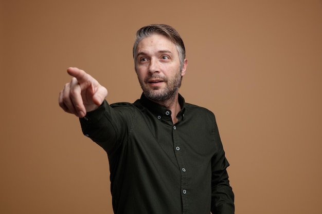 Mann mittleren Alters mit grauem Haar in dunklem Hemd, der lächelnd zur Seite schaut und mit dem Zeigefinger auf die Seite zeigt, die vor braunem Hintergrund steht