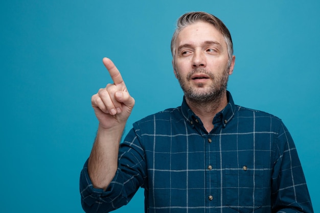 Kostenloses Foto mann mittleren alters mit grauem haar in dunklem hemd, der schlau zur seite schaut und mit dem zeigefinger auf etwas zeigt, das über blauem hintergrund steht