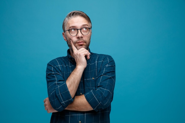 Mann mittleren Alters mit grauem Haar in dunklem Hemd mit Brille, der verwirrt nach oben blickt und über blauem Hintergrund steht