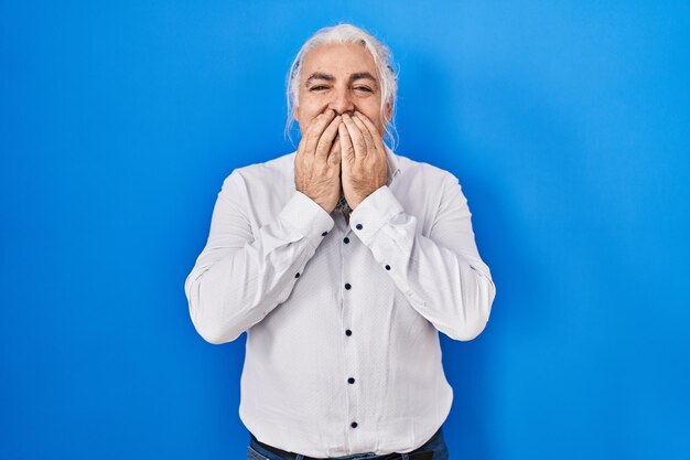 Mann mittleren Alters mit grauen Haaren, der vor blauem Hintergrund steht und lachend und verlegen kichert und den Mund mit den Händen bedeckt, Klatsch und Skandal-Konzept
